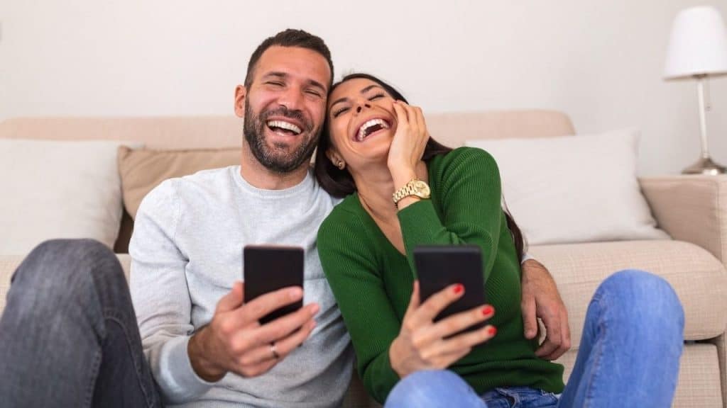 A man and woman sit together laughing while holding their respective smartphones.