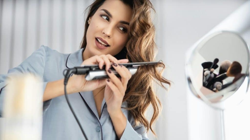 A woman styles her hair with a flat iron while looking into a vanity mirror.