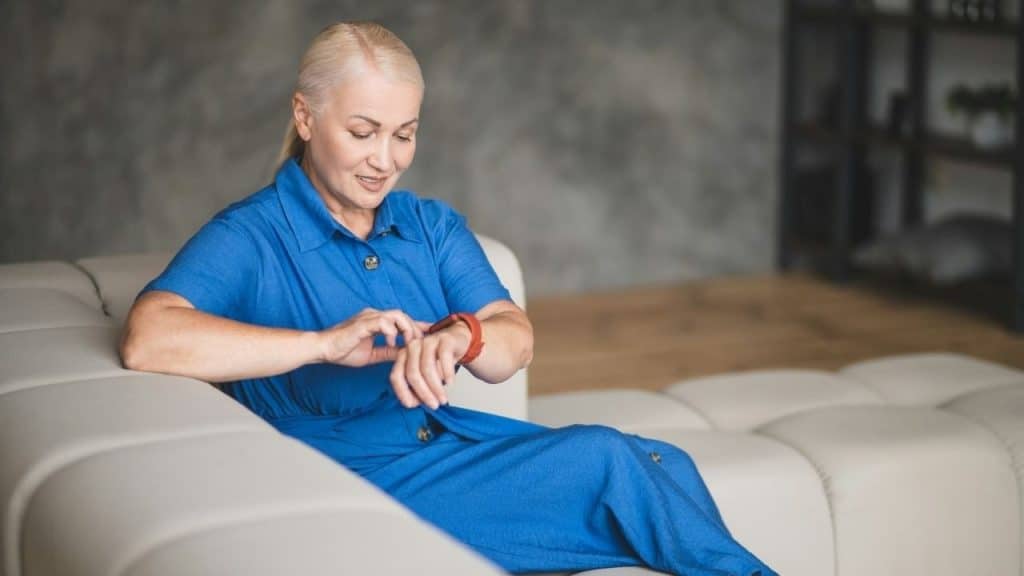 A woman in a blue dress sits on a sofa and adjusts her red smartwatch.