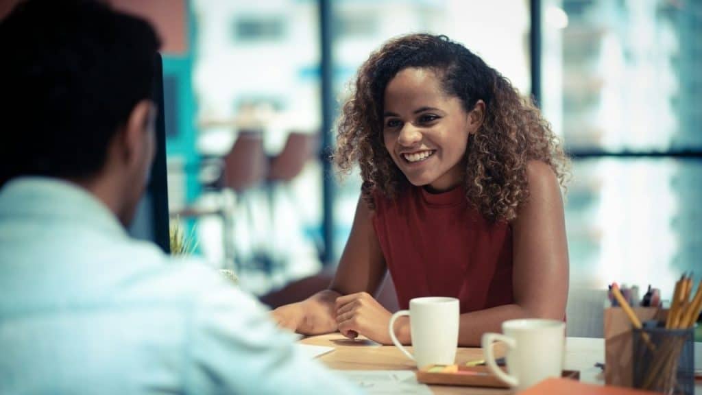 A smiling woman with curly hair looks at a man while sitting at a desk.