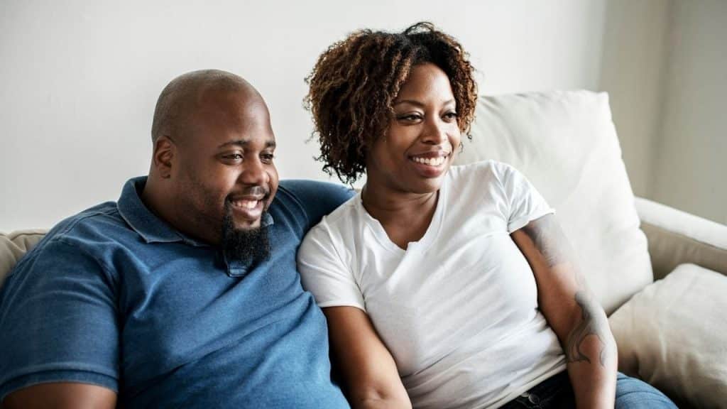 A man and woman sit close together on a light-colored couch, both smiling.