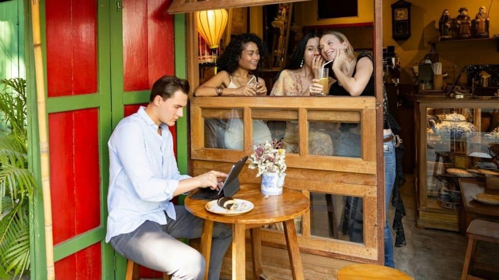 Three women whisper and smile while looking at a man working on a tablet nearby.