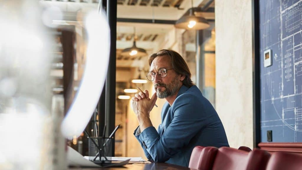 A thoughtful man with glasses sits at a desk in a modern office.