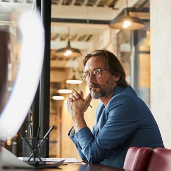 A thoughtful man with glasses sits at a desk in a modern office.