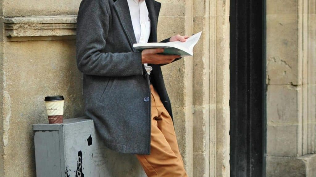 A person leaning against a stone wall while reading a book beside a takeaway coffee cup.