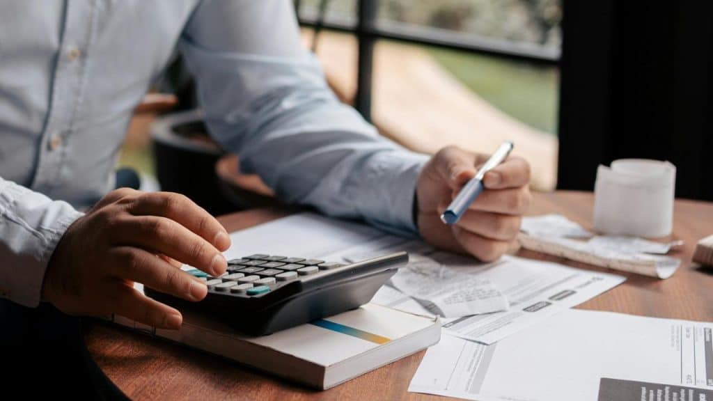 A person using a calculator and pen while reviewing bills and paperwork at a desk.
