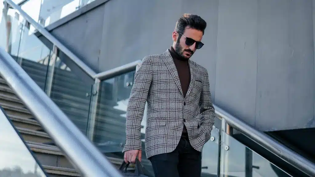 A stylish man wearing sunglasses and a plaid blazer walking down modern outdoor stairs.