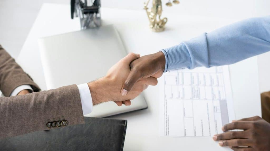 A business handshake taking place over a desk with documents and a laptop.