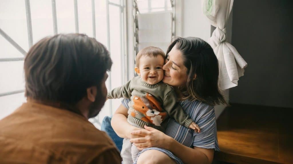 A smiling baby held by a parent while another parent looks on by a window.