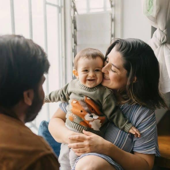 A smiling baby held by a parent while another parent looks on by a window.