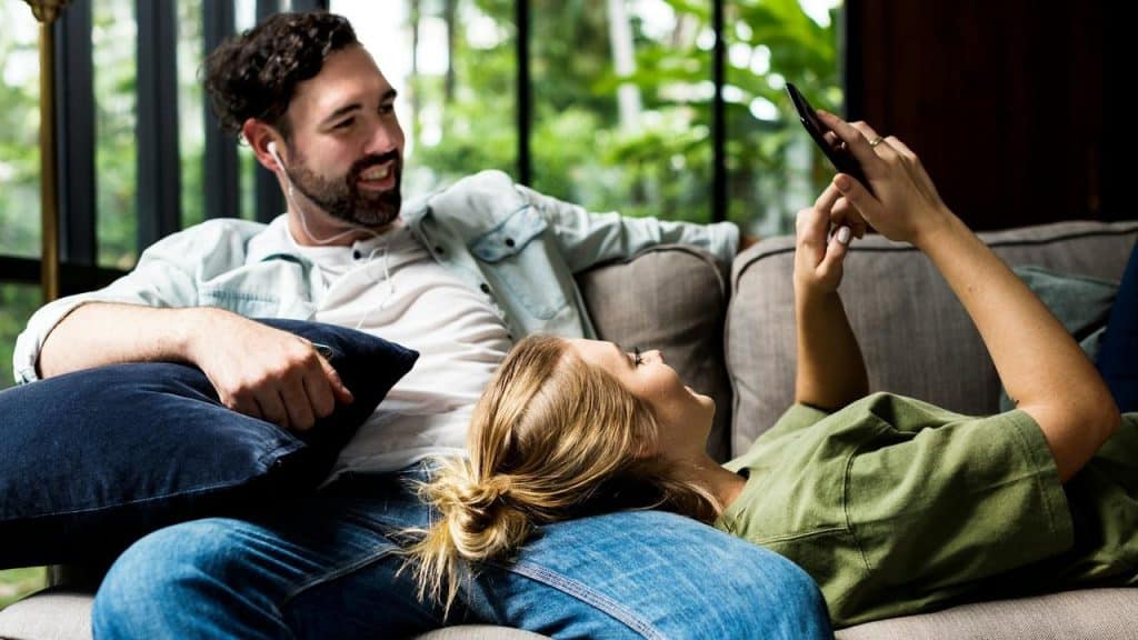 A couple relaxing on a couch while one person looks at a smartphone.