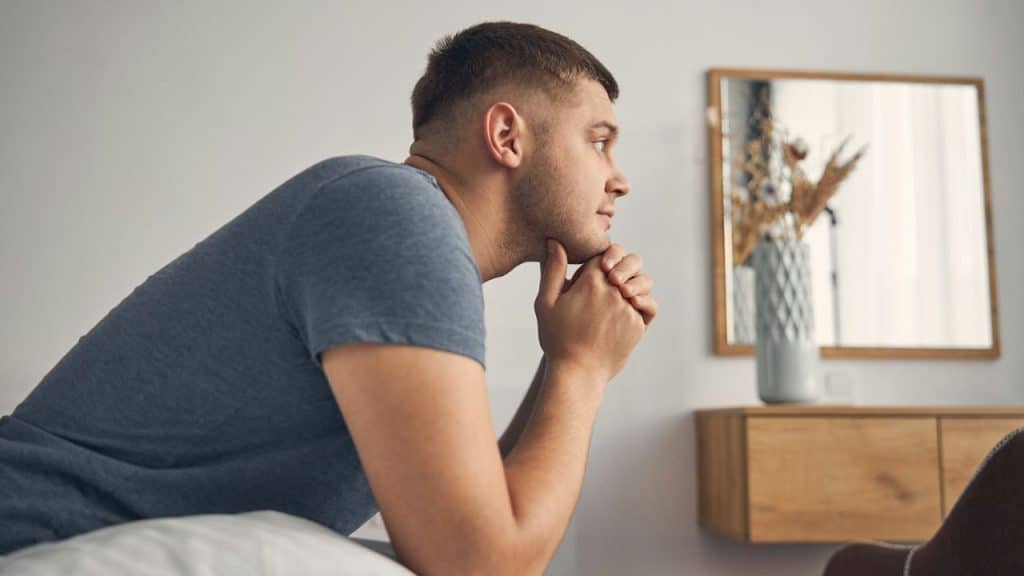 Man with a short haircut sitting on a bed or sofa, resting his chin on his clasped hands.