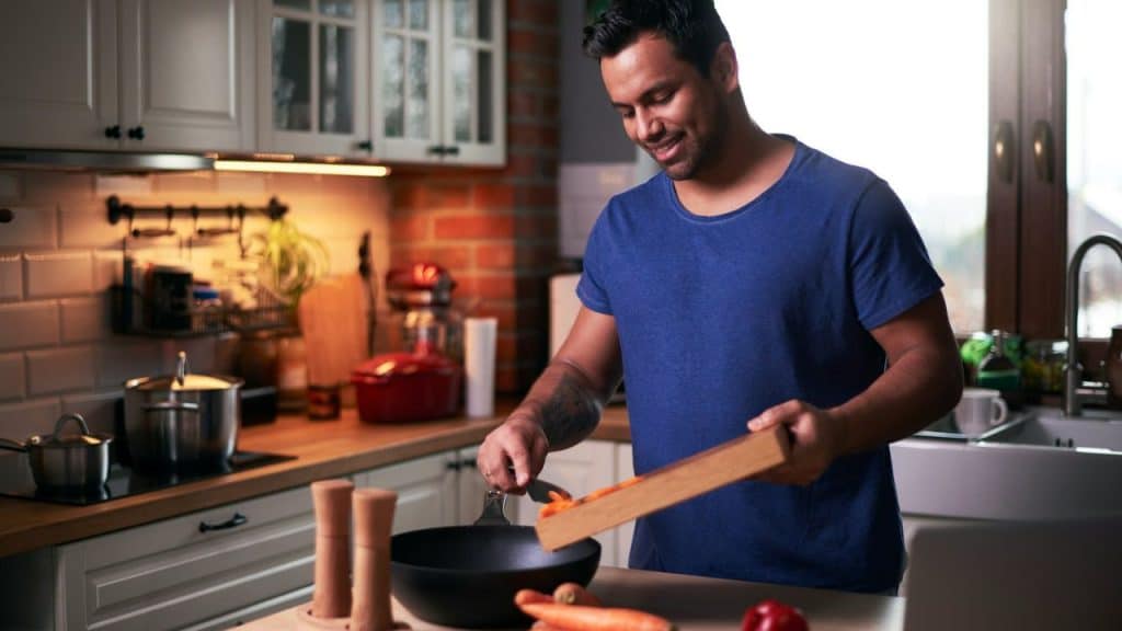 Smiling man in a blue shirt adding chopped carrots from a cutting board into a pan in a kitchen.