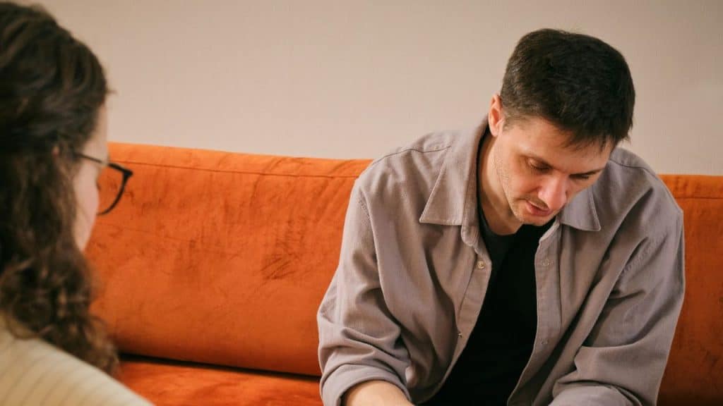 Man sitting on an orange couch looking down while a person with glasses talks to him.