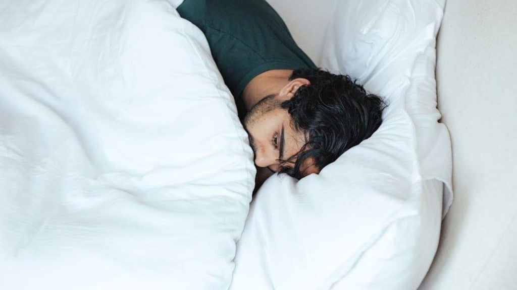 Man with dark hair lying in bed under white covers, turned away from the camera.