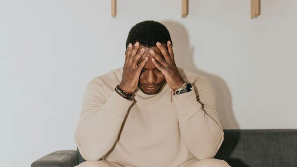 Man wearing a beige sweatshirt sitting on a sofa with his hands clasped over his head.