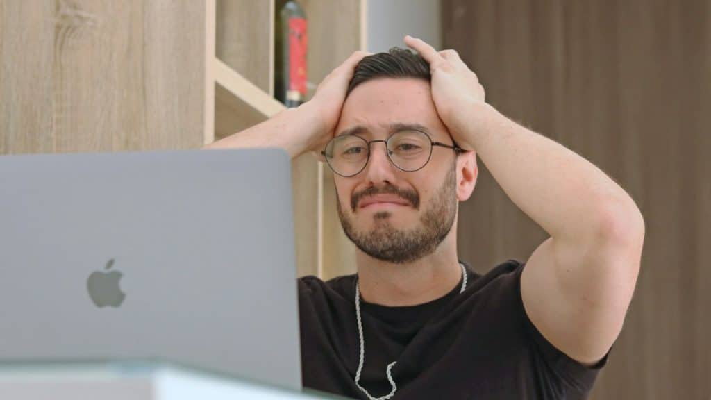Frustrated man with glasses, a beard, and a silver necklace holding his head in his hands in front of a laptop.