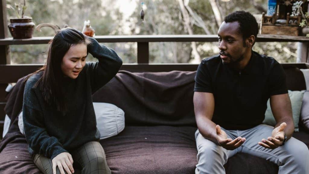 Two people sitting far apart on an outdoor sofa, a woman looking upset and a man talking with open hands.