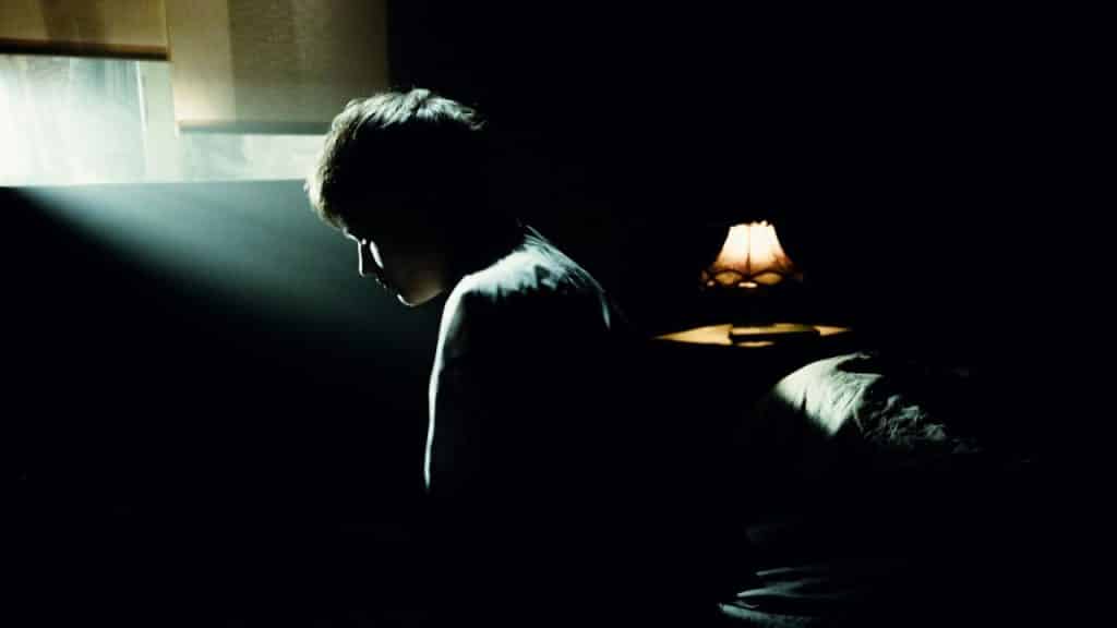 Person sitting alone in a darkened room, illuminated by a spotlight from a window and a bedside lamp.