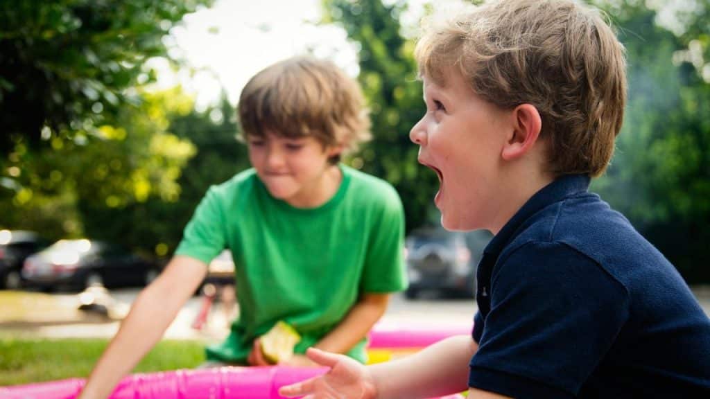 A boy laughing in a small inflatable pool with a friend outdoors.