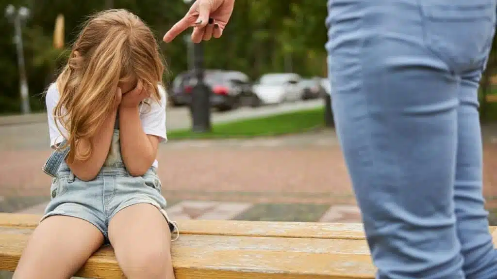 A girl on a bench covering her face as an adult points at her.