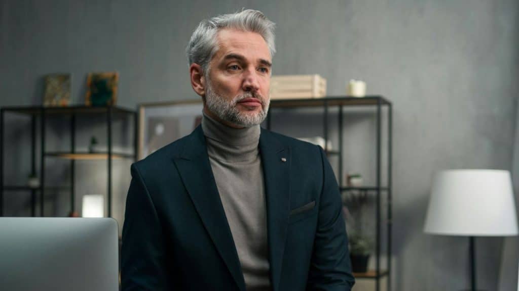 A silver-haired, bearded man in a suit jacket and turtleneck sits in a modern office.