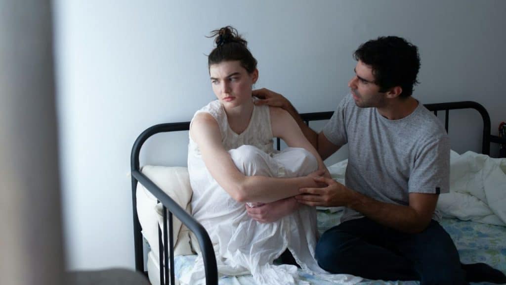 A man comforts a distressed woman sitting on a metal-framed bed with white walls.
