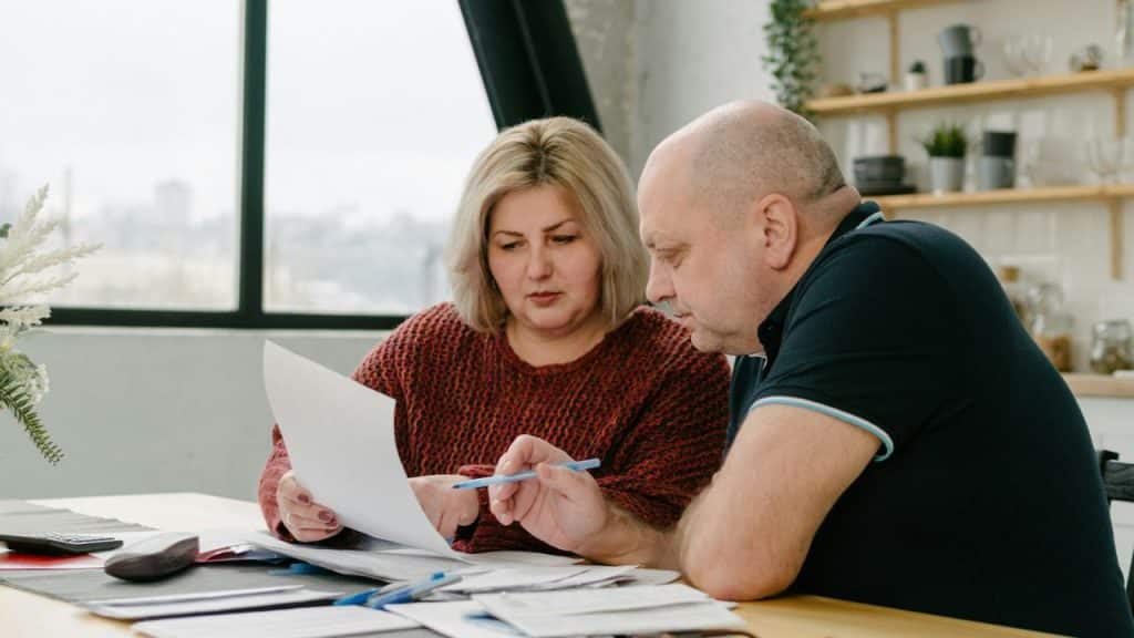 A blonde woman and a bald man review papers together at a table in a bright kitchen.