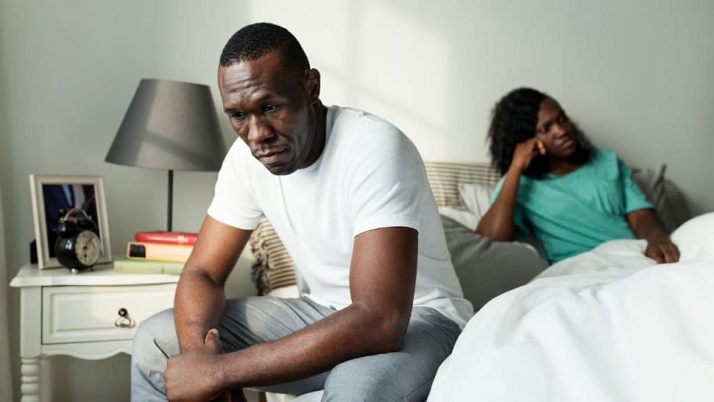 A somber man sits on the edge of a bed while a woman sits behind him, looking away.