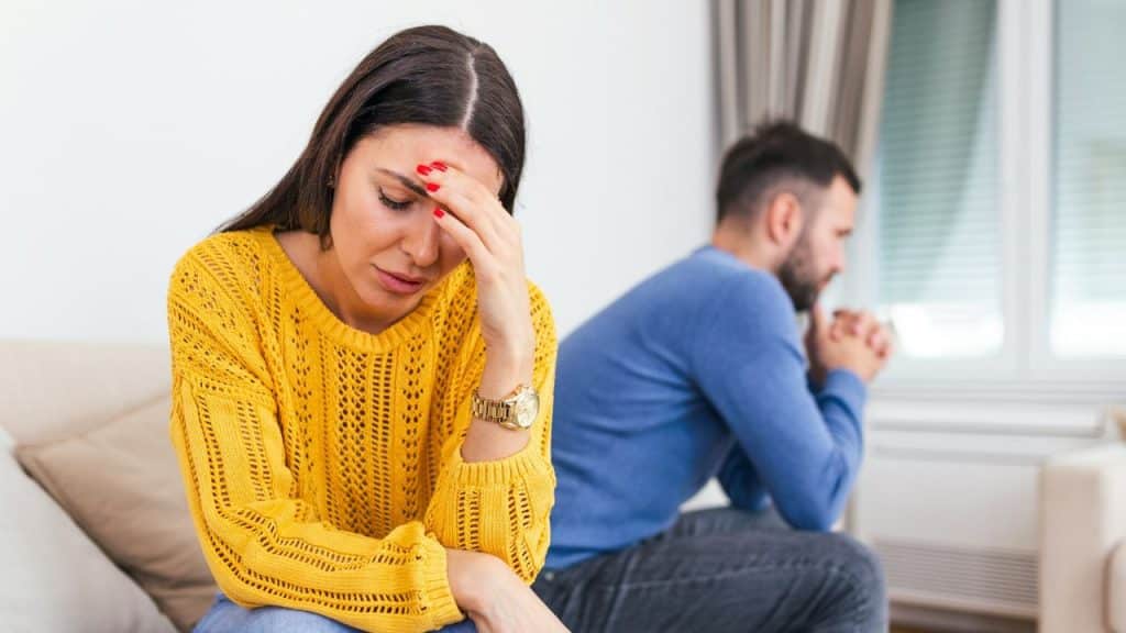 A distressed woman with her hand on her forehead sits in front of a man looking away.