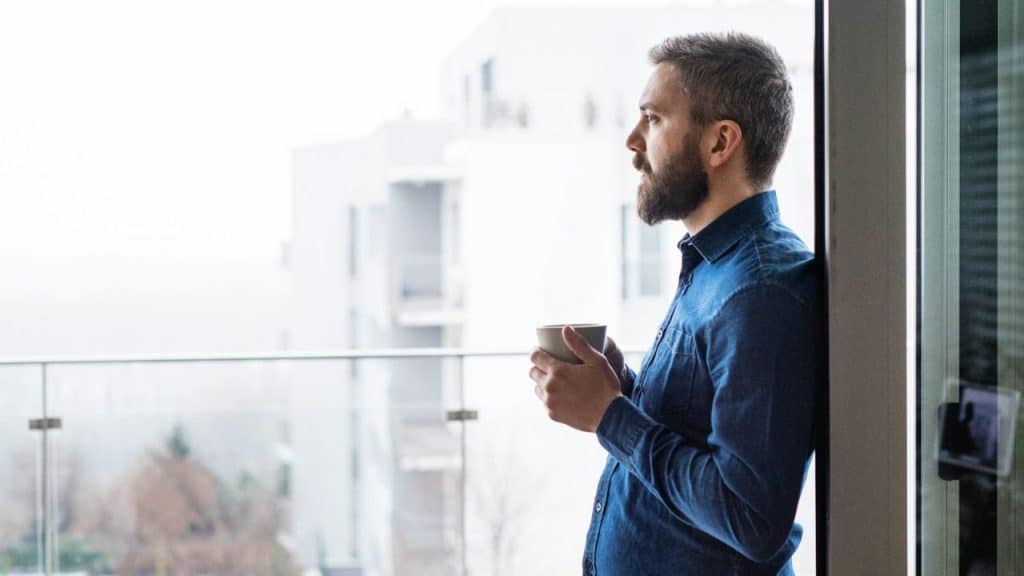 A bearded man leans on a glass door, holding a mug and looking at an apartment complex.