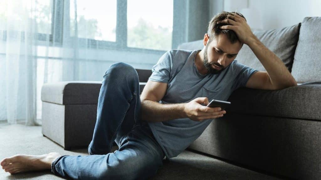 A stressed man sits on the floor holding his head in one hand and looking at his smartphone in the other.
