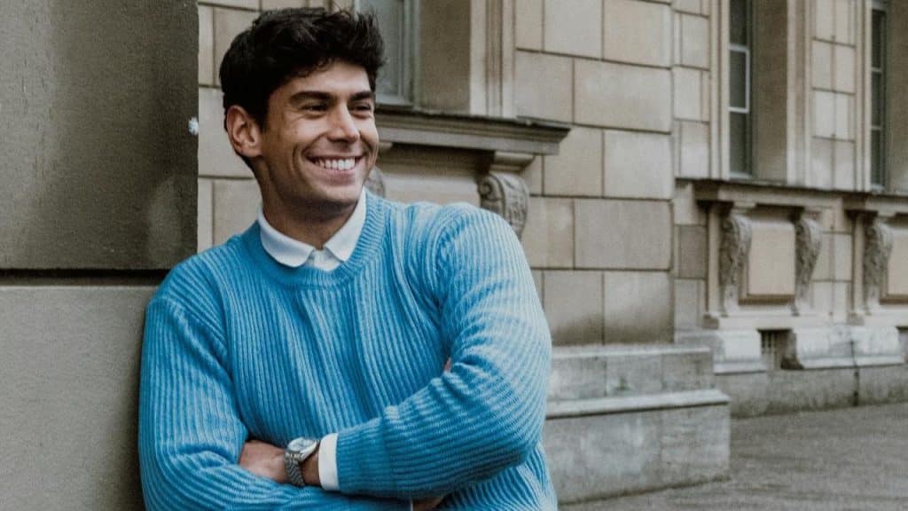 Smiling man in a blue sweater leaning against a stone building with his arms crossed.