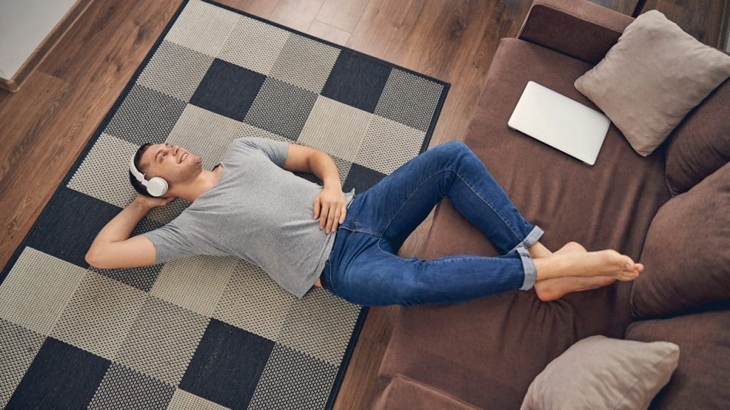 A man wearing headphones is lying on the floor with his feet resting on a couch and listening to music.