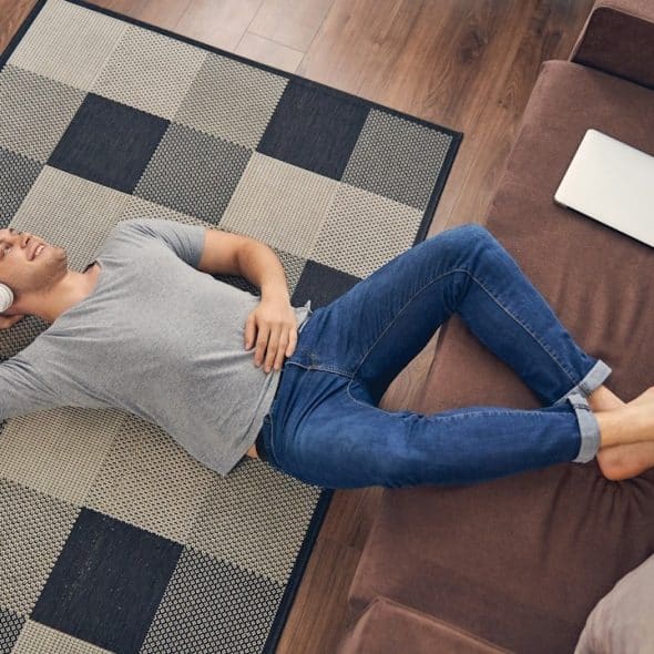 A man wearing headphones is lying on the floor with his feet resting on a couch and listening to music.