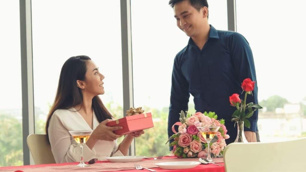 A man presents a gift to a woman seated at a table with roses and wine.