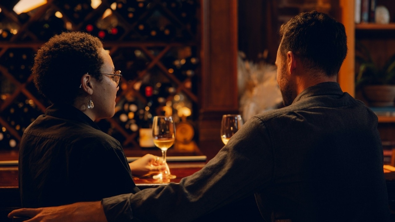 A man and a woman sit at a bar with wine glasses in front of them.