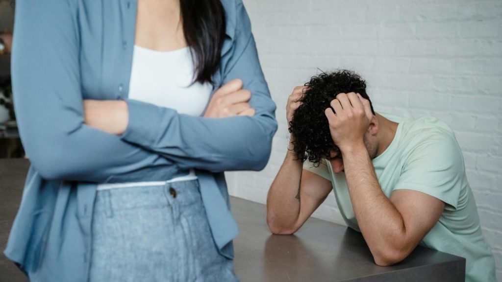 A man sits with his head in his hands near a woman with crossed arms.