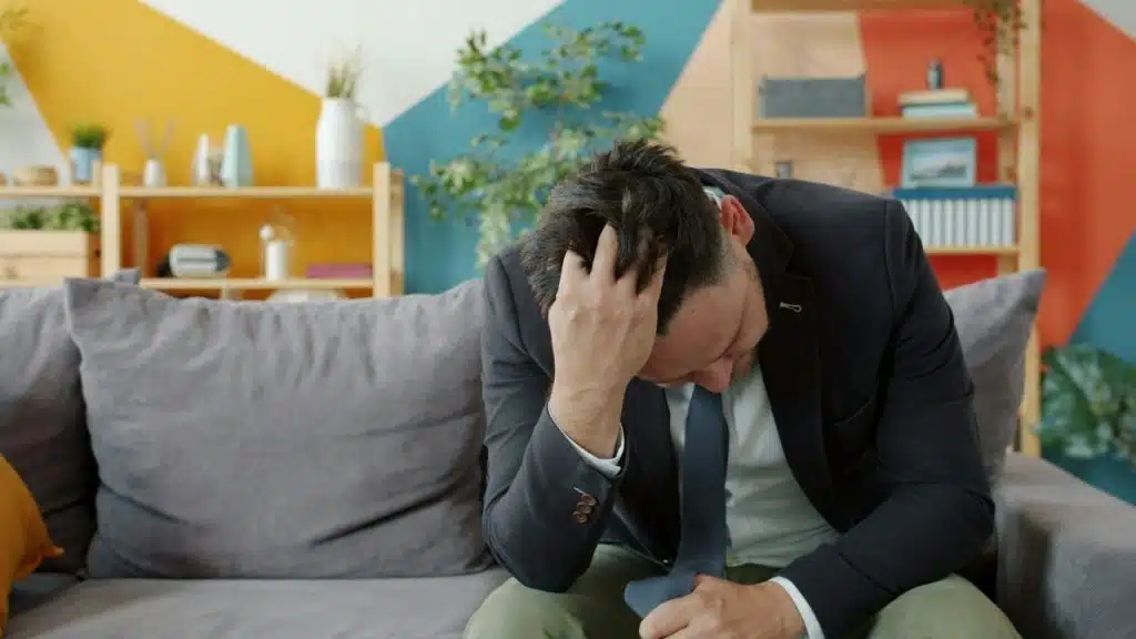 A man in a suit sits on a gray sofa with his hand on his head.