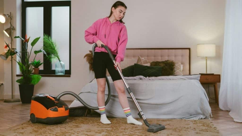 A woman in a pink hoodie vacuums a rug in a bedroom with an orange vacuum.