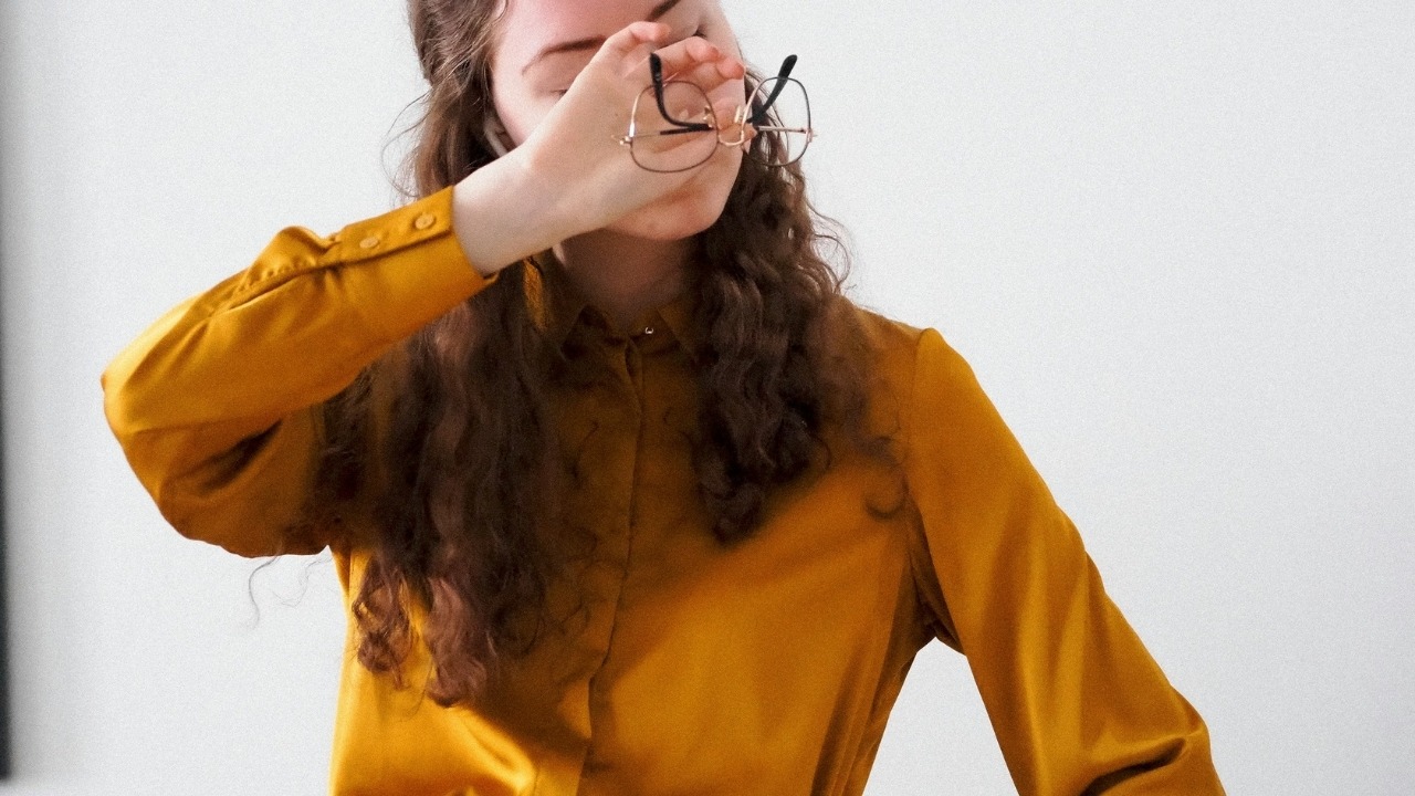 A woman in a gold shirt holds her glasses while covering her eyes with her hand.