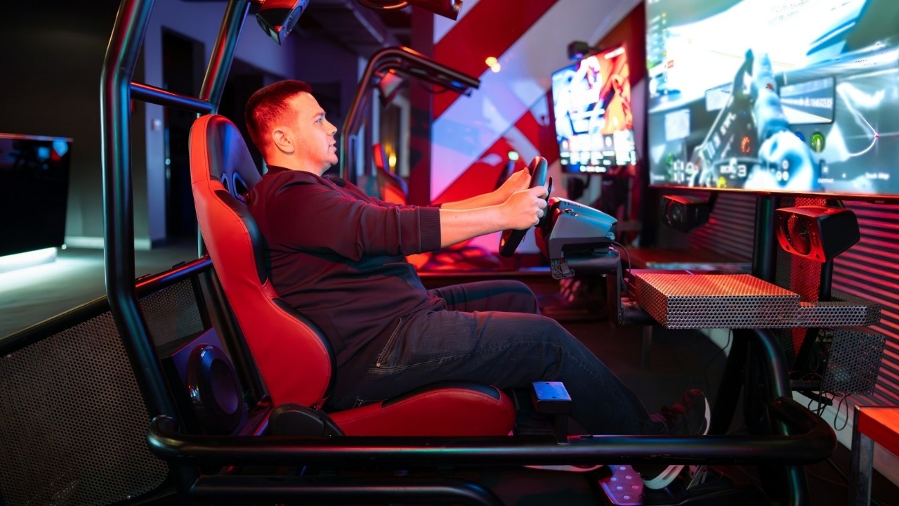 A man sits in a red racing simulator chair playing a video game with a wheel.
