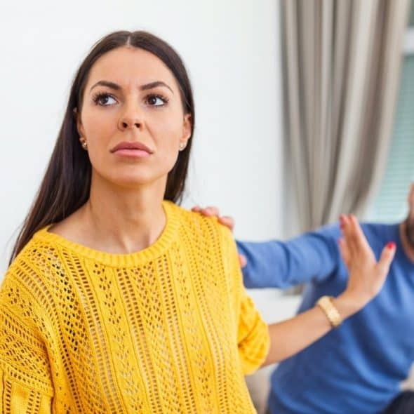 A woman in a yellow sweater looks away while a man gestures toward her behind.