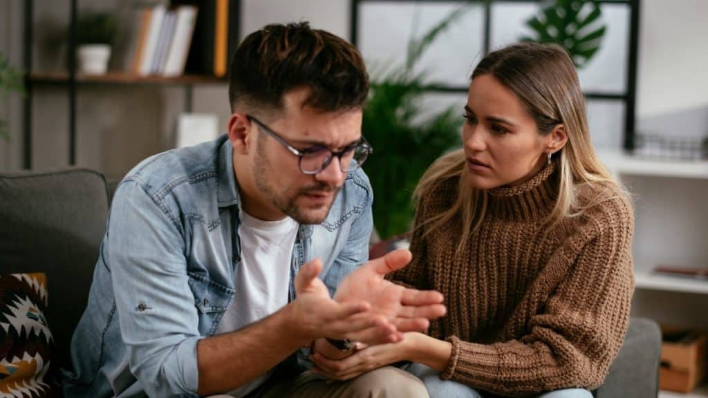 A man with glasses gestures with his hands while a woman in a brown sweater listens.