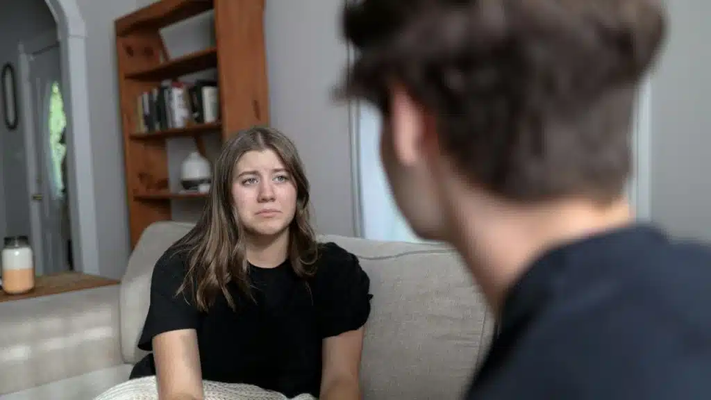 A young woman with a sad expression sits on a couch looking at a man.