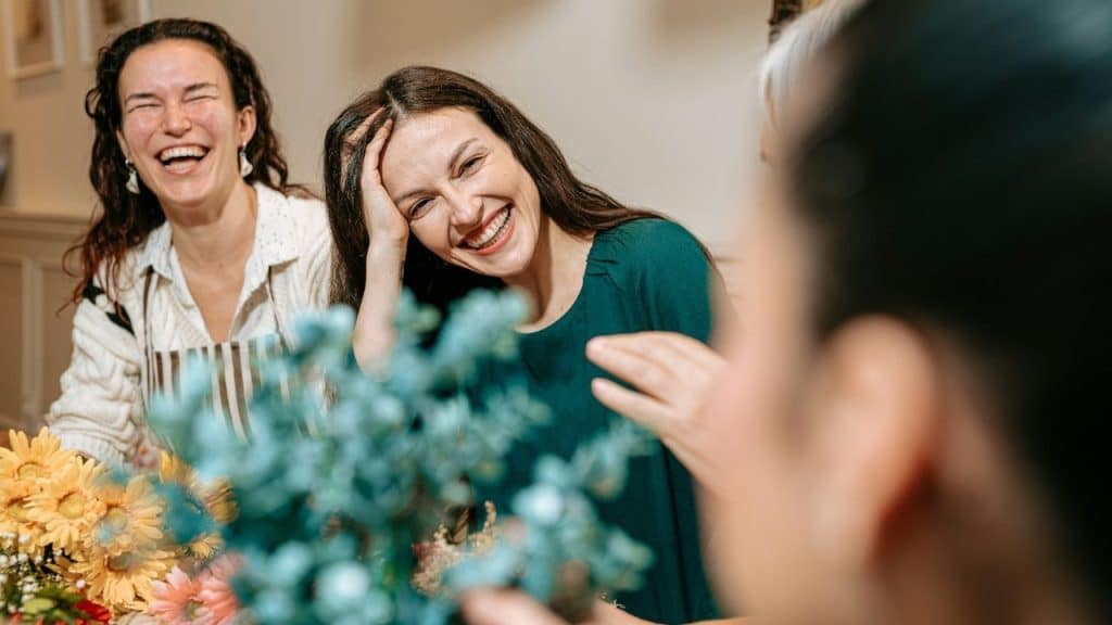 Three women are laughing together while sitting around a table with various colorful flowers.