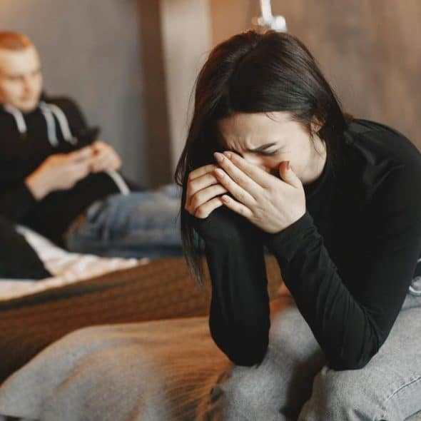 A woman in black sits crying into her hands while a man sits blurred behind her.