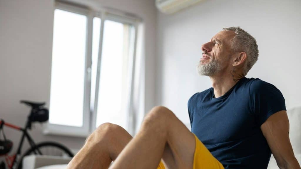 A man with grey hair and a beard sits indoors and looks toward a window.