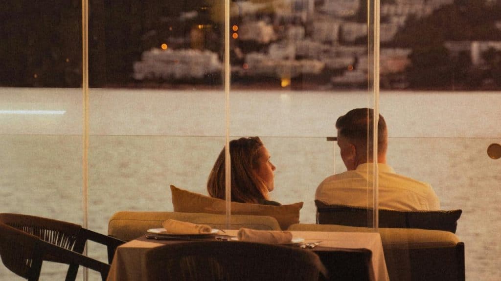 A couple sitting at a waterfront restaurant, talking through a glass wall at dusk.