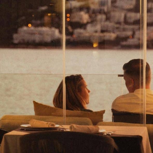 A couple sitting at a waterfront restaurant, talking through a glass wall at dusk.